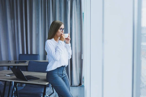 Young smiling business woman standing in office near window with coffee in hands. A female manager in glasses and a white shirt is resting from work