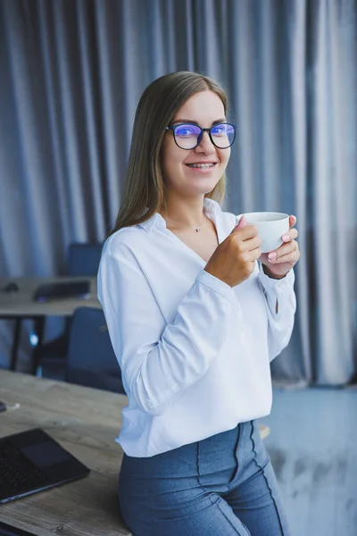 Young smiling business woman standing in office near window with coffee in hands. A female manager in glasses and a white shirt is resting from work