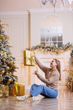 Genç ve mutlu bir kadın yeni yıl hediyesiyle oturuyor. Noel ağacı ve süslü çelenkleri olan bir şömine. Yeni yıl havası.
