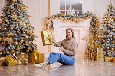 Genç ve mutlu bir kadın yeni yıl hediyesiyle oturuyor. Noel ağacı ve süslü çelenkleri olan bir şömine. Yeni yıl havası.