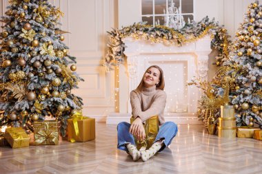 Genç ve mutlu bir kadın yeni yıl hediyesiyle oturuyor. Noel ağacı ve süslü çelenkleri olan bir şömine. Yeni yıl havası.