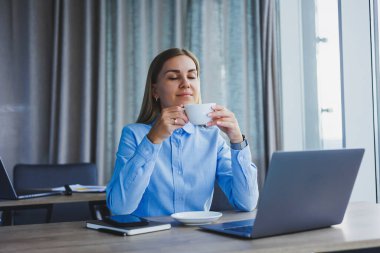 Portrait of cheerful woman in classic glasses smiling in free time in cafe with coffee, positive european woman in blue shirt, desk with laptop, remote work