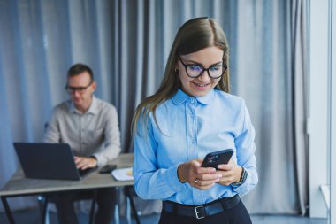Manager woman in classic glasses smiling during working hours in the office talking on the phone, table with laptop, colleagues in the background. A colleague is in the background, selective focus