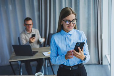 Manager woman in classic glasses smiling during working hours in the office talking on the phone, table with laptop, colleagues in the background. A colleague is in the background, selective focus