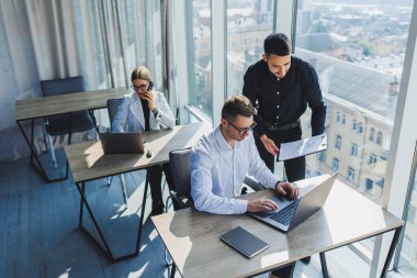 Gözlüklü başarılı insanlar tahta bir masada dizüstü bilgisayarıyla oturup modern bir ofiste çalışıyorlar..