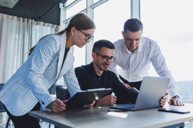 Resmi olarak bir grup iş adamı proje detaylarını tartışıyor ve modern bir toplantı odasında iş birliği yaparken dizüstü bilgisayar ekranına bakıyor. Modern ofis