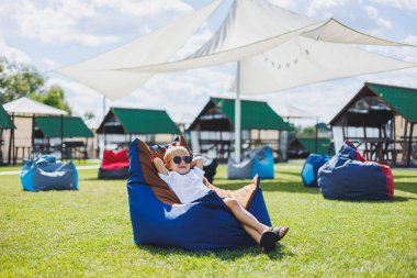 Beyaz tişörtlü ve şortlu, 5-6 yaşlarında açık renk saçlı bir çocuk fasulye koltukta oturuyor. Güneş gözlüklü bir çocuk temiz havada dinleniyor.
