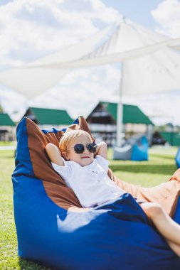 Beyaz tişörtlü ve şortlu, 5-6 yaşlarında açık renk saçlı bir çocuk fasulye koltukta oturuyor. Güneş gözlüklü bir çocuk temiz havada dinleniyor.