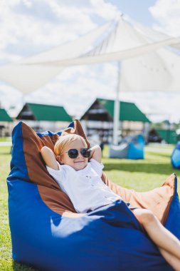 Beyaz tişörtlü ve şortlu, 5-6 yaşlarında açık renk saçlı bir çocuk fasulye koltukta oturuyor. Güneş gözlüklü bir çocuk temiz havada dinleniyor.