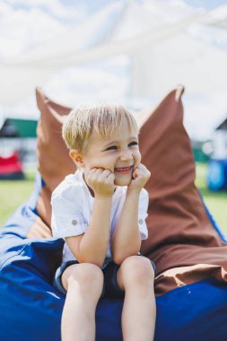 Mutlu çocuk yaz parkında koltukta oturuyor. Beyaz tişörtlü küçük bir çocuk dışarıda eğleniyor. Çocukluk ve mutluluk.