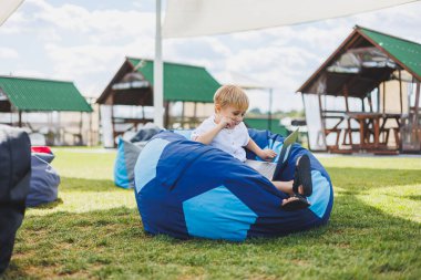 Yaz çimlerinin arkasında dizüstü bilgisayarı olan küçük bir çocuk. Beyaz tişörtlü bir çocuk parkta bir armut çuvalının üzerinde oturuyor..