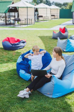 Mutlu oğul ve anne doğada oturup dizüstü bilgisayara bakıyor. Genç bir annenin uzaktan kumandası. Aile tatili