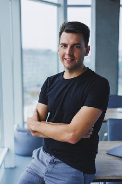 Modern bir ofisin arka planında siyah tişörtlü gülümseyen genç bir adamın portresi. Günlük kıyafetlerle şık bir adam.