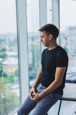 Modern bir ofisin arka planında siyah tişörtlü gülümseyen genç bir adamın portresi. Günlük kıyafetlerle şık bir adam.