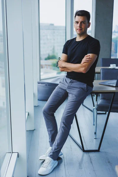 Portrait of a smiling young man in a black t-shirt against the background of a modern office. Stylish guy in casual clothes