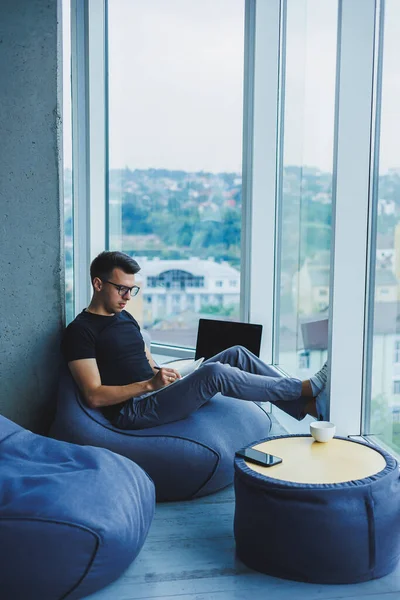 A young male student of Caucasian appearance sits in a bag chair and takes notes in a notebook. The manager sits by the window and writes the work schedule