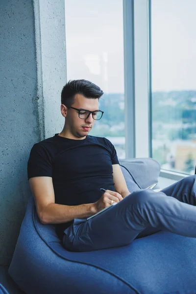 A young male student of Caucasian appearance sits in a bag chair and takes notes in a notebook. The manager sits by the window and writes the work schedule