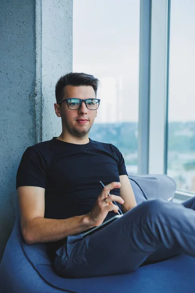 A young male student of Caucasian appearance sits in a bag chair and takes notes in a notebook. The manager sits by the window and writes the work schedule