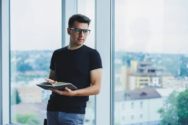 A student manager stands near a large window and reads notes in a notebook. A young man in a black T-shirt with glasses stands in the office near the window