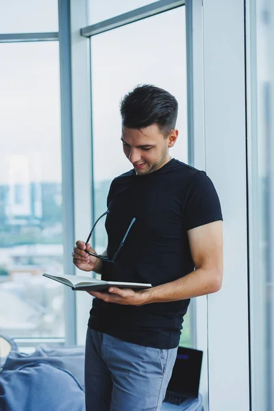 A student manager stands near a large window and reads notes in a notebook. A young man in a black T-shirt with glasses stands in the office near the window