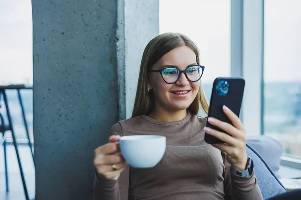 Blonde young smiling woman in glasses in casual clothes sitting in a chair by the window with a phone and drinking coffee