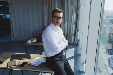 Gözlüklü ve beyaz gömlekli sevimli, vahşi iş adamı, modern ofiste büyük pencereleri var. Klasik pantolonlu ve beyaz gömlekli bir adam. Genç çekici iş adamı