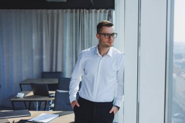 Gözlüklü ve beyaz gömlekli sevimli, vahşi iş adamı, modern ofiste büyük pencereleri var. Klasik pantolonlu ve beyaz gömlekli bir adam. Genç çekici iş adamı