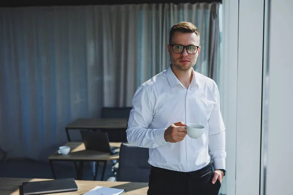 Cute young businessman with coffee in hand in glasses and white shirt, he is in modern office with big windows. A man in classic trousers and a white shirt. Young attractive businessman