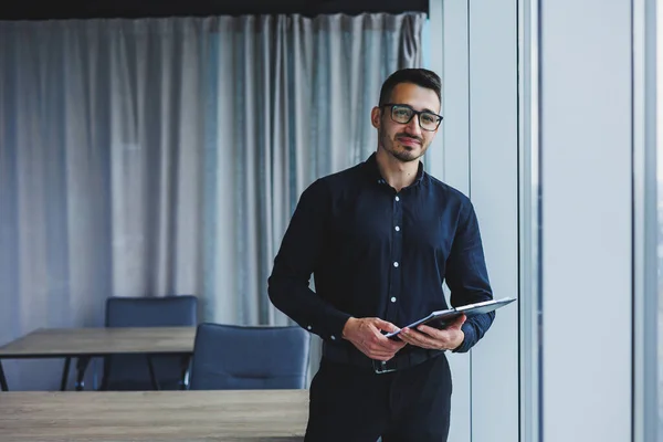 A Caucasian business man in glasses and a black shirt sits in the office and looks at sales growth charts. A young successful businessman in classic clothes stands by the window in the office