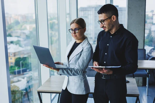 Beautiful businesswoman standing with Caucasian male colleague watching laptop screen and talking with businessman in office. Business relations between colleagues