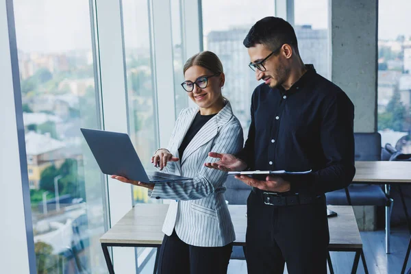 Beautiful businesswoman standing with Caucasian male colleague watching laptop screen and talking with businessman in office. Business relations between colleagues