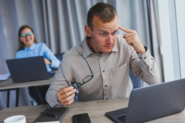 Dissatisfied businessman looking at something on smartphone while working. The concept of a modern successful person. Young confused guy in glasses sitting at desk in open space office with laptops