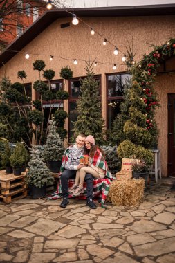 İskoç battaniyesine sarılı güzel, mutlu, genç bir çift. Arka planda yeşil Noel ağaçları var. Kış tatili. Yeni yılı bekliyorum.