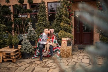 İskoç battaniyesine sarılı güzel, mutlu, genç bir çift. Arka planda yeşil Noel ağaçları var. Kış tatili. Yeni yılı bekliyorum.