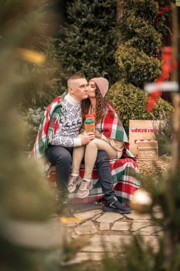 İskoç battaniyesine sarılı güzel, mutlu, genç bir çift. Arka planda yeşil Noel ağaçları var. Kış tatili. Yeni yılı bekliyorum.