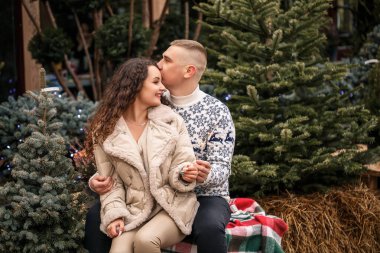 Noel ağaçlarının yanında beyaz elbiseli, çelenklerin yanında yürüyen güzel bir çift. Mutlu erkek ve kadın, romantizm, Noel kutlaması, eğlence, aşk. Beyaz süveter, yeni yıl, sokak, dışarıda