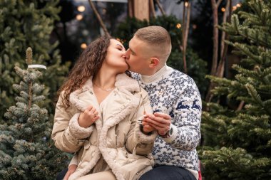 Noel ağaçlarının yanında beyaz elbiseli, çelenklerin yanında yürüyen güzel bir çift. Mutlu erkek ve kadın, romantizm, Noel kutlaması, eğlence, aşk. Beyaz süveter, yeni yıl, sokak, dışarıda