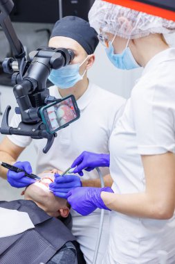 Genç bir dişçi diş mikroskobuyla hastanın dişlerine bakar ve diş aletlerini ağzının yakınında tutar. Bir asistan doktora yardım eder. Maskeli beyaz üniformalar giyiyorlar.