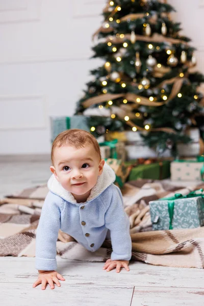 A one-year-old boy sits near a decorated Christmas tree with gifts. Christmas tree in the house. A happy child is waiting for the new year