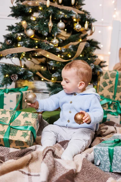 A one-year-old boy sits near a decorated Christmas tree with gifts. Christmas tree in the house. A happy child is waiting for the new year