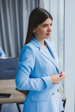 Ofisteki panoramik pencerenin yanında şehir manzaralı mavi ceketli güzel bir esmerin portresi. Ofisteki mutlu iş kadını.