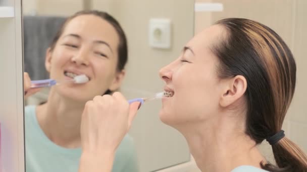 Mujer con aparatos ortopédicos limpia sus dientes con un cepillo de dientes, ante el espejo, vista superior. — Vídeos de Stock