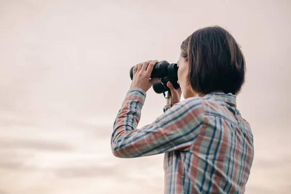 Ung oigenkännlig kvinna tittar genom kikare i bakgrunden av himlen. — Stockfoto