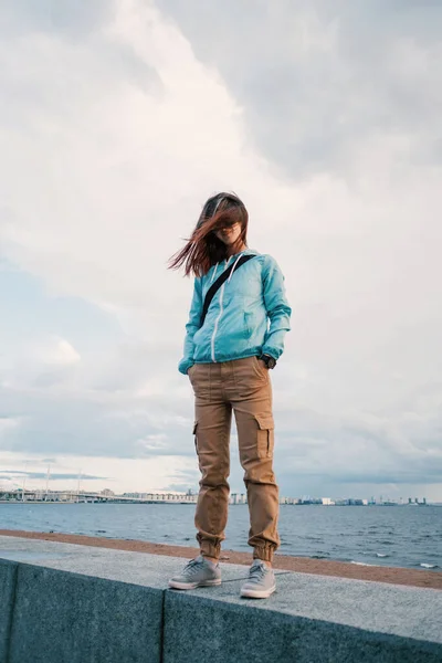 A full-length portrait of a young woman in a blue windbreaker in windy weather. — Stock Photo, Image