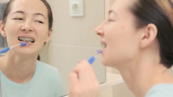 Mujer joven con aparatos ortopédicos limpia sus dientes con un cepillo dental especial, ante el espejo. — Vídeos de Stock