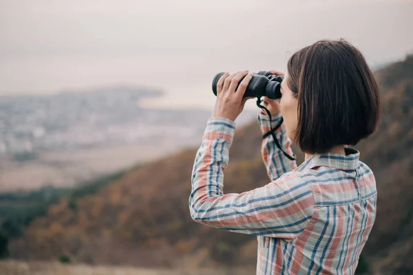 Mujer joven mira a través de los prismáticos y admira el paisaje de montaña. — Foto de Stock