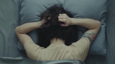Yatakta yastığa gömülü bir kadın ellerini başına bastırıyor..