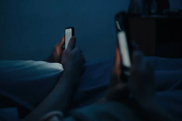 Seorang pria dan wanita berbaring di pastel di malam hari dan menggunakan ponsel mereka. Memeriksa media sosial sebelum tidur. — Stok Foto