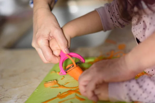 Little cute baby toddler girl in the kitchen peeling carrots with carrot peeler on chopping board. Child help at home, closeup hands. — Stock Photo, Image