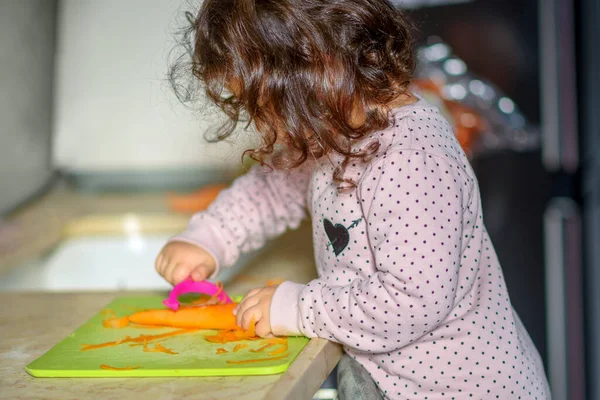 Маленькая милая малышка на кухне, чистит морковь с кожурой для моркови на доске. Помощь детям на дому, крупный план. — стоковое фото